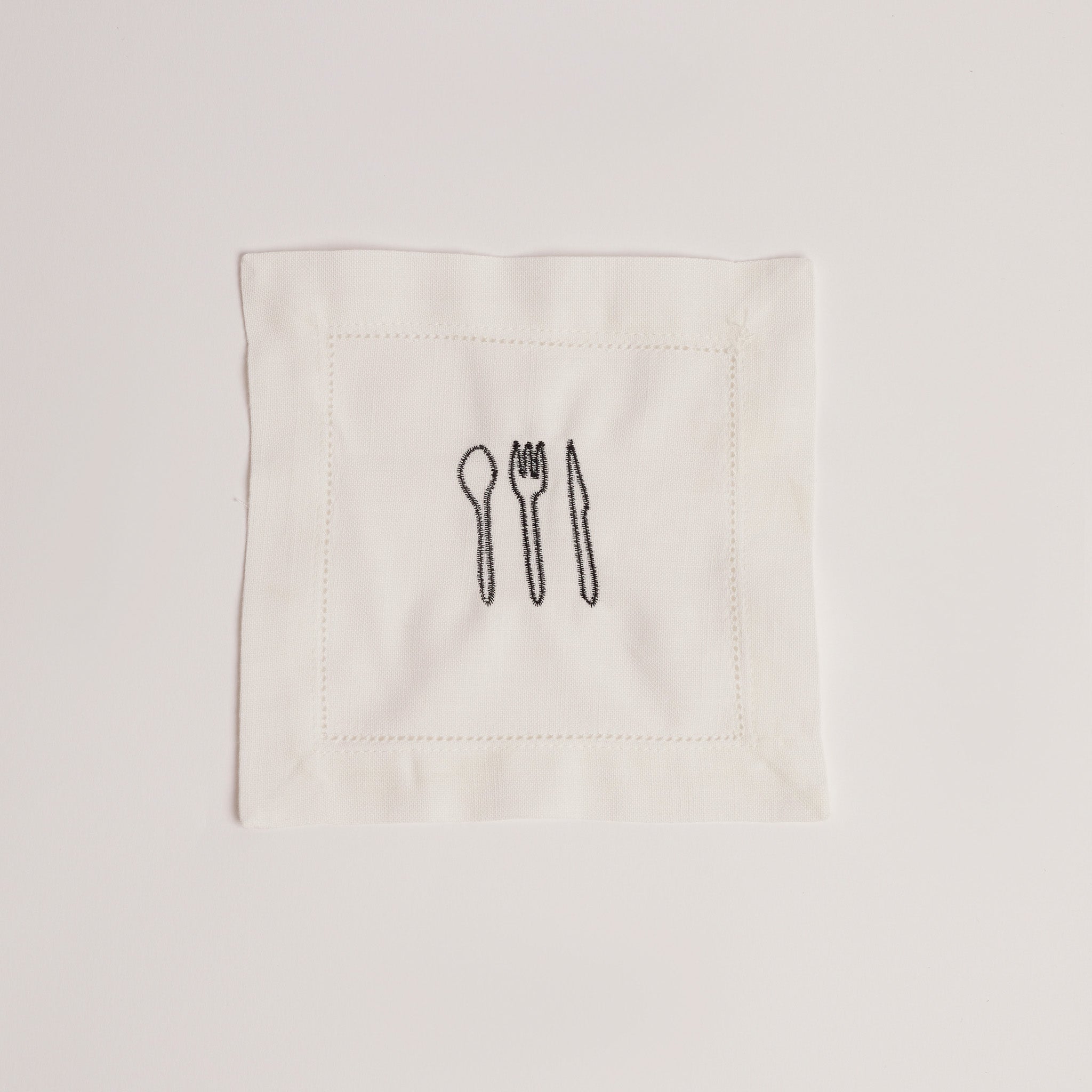 Single white cocktail napkin embroidered with black cutlery motif. Single white cocktail napkin embroidered with black cutlery motif.