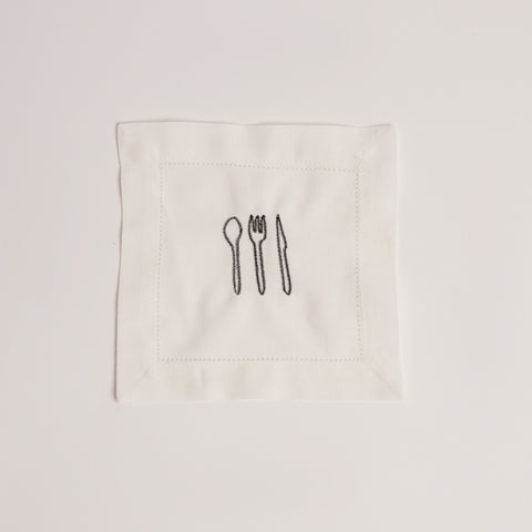 Single white cocktail napkin embroidered with black cutlery motif. Single white cocktail napkin embroidered with black cutlery motif.