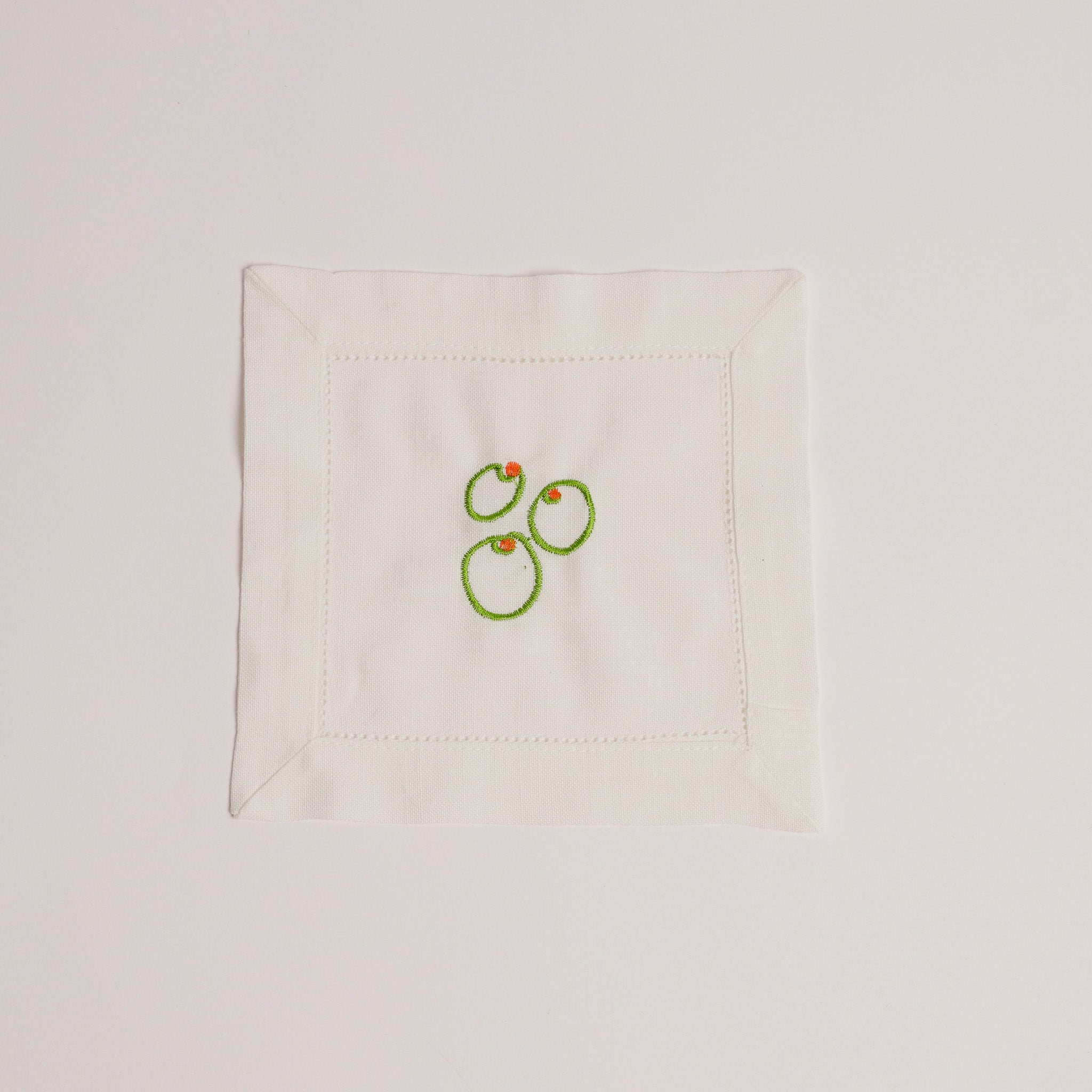Single white cocktail napkin embroidered with three green olives and red pimento detail. Single white cocktail napkin embroidered with three green olives and red pimento detail.