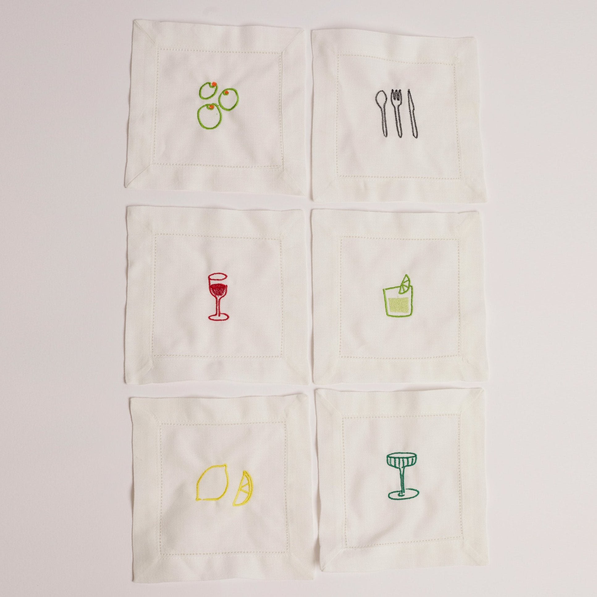 A set of white cocktail napkins with various embroidered designs including a glass of red wine, a lemon, an orange, a cucumber, and a cocktail glass. A set of white cocktail napkins with various embroidered designs including a glass of red wine, a lemon, an orange, a cucumber, and a cocktail glass.