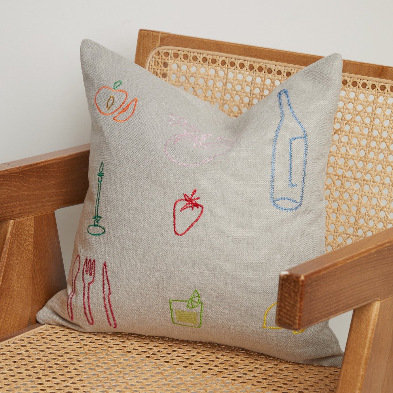 Close-up of neutral linen cushion with colourful embroidered fruit, cutlery, and bottle icons on a cane wooden chair. Close-up of neutral linen cushion with colourful embroidered fruit, cutlery, and bottle icons on a cane wooden chair.