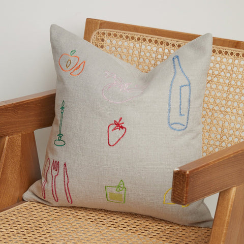 Close-up of neutral linen cushion with colourful embroidered fruit, cutlery, and bottle icons on a cane wooden chair. Close-up of neutral linen cushion with colourful embroidered fruit, cutlery, and bottle icons on a cane wooden chair.