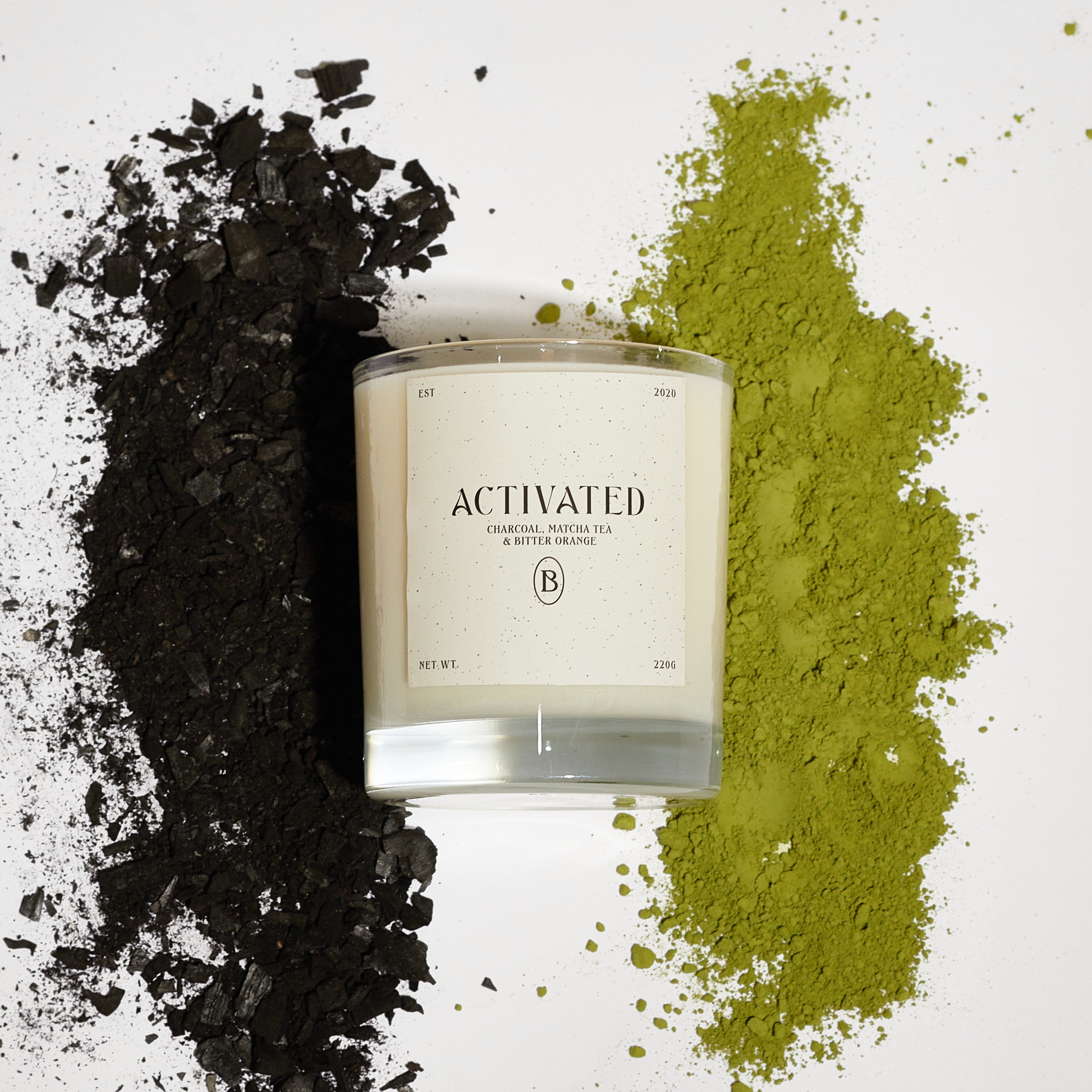 Boujie Activated jar candle in a sleek clear glass jar, styled in a flatlay with crushed charcoal on one side and vibrant green matcha powder on the other, capturing the bold, uplifting scent of charcoal, matcha tea, and bitter orange.