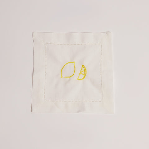 Single white cocktail napkin embroidered with a yellow lemon and lemon wedge.