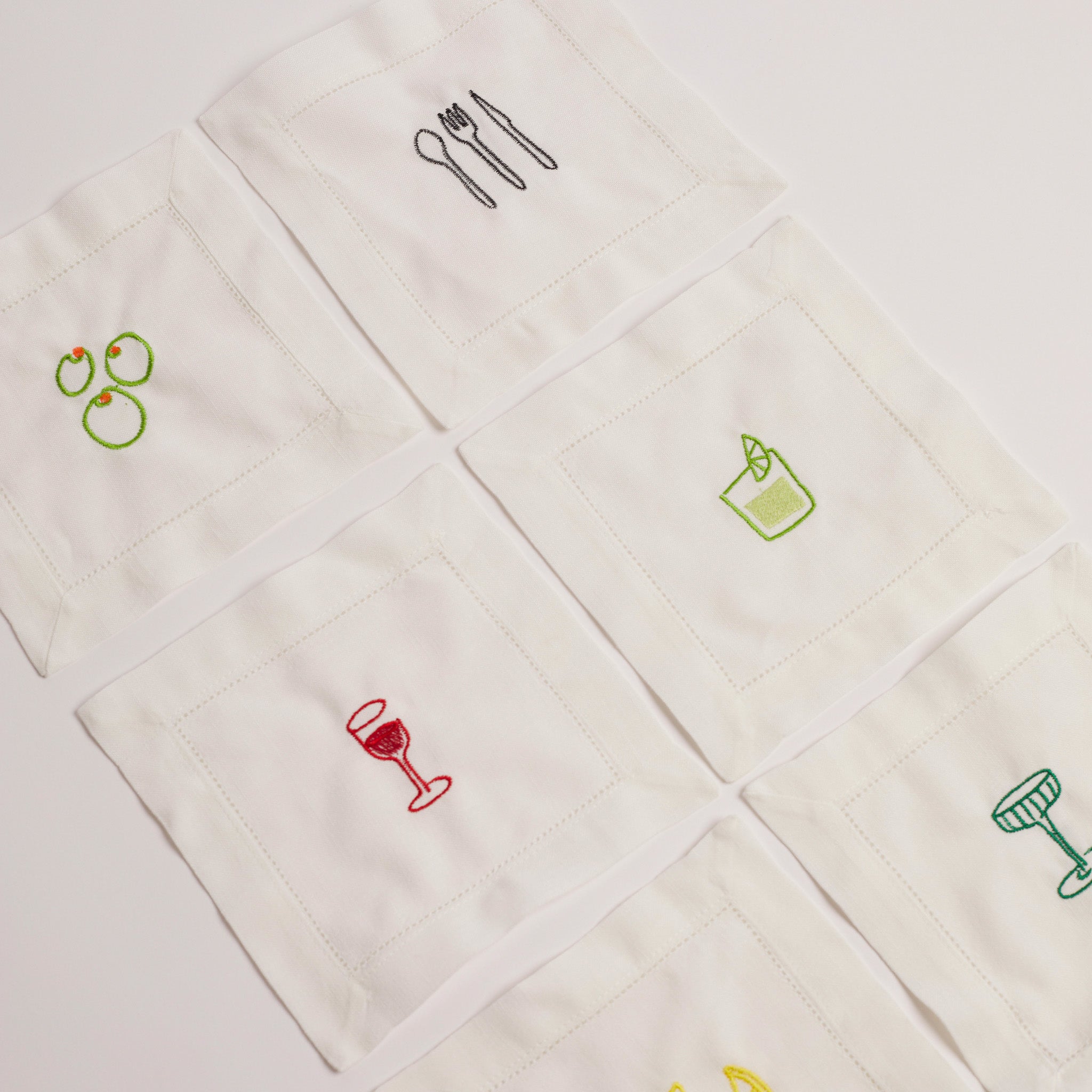 Full set of embroidered cocktail napkins laid flat, showing six colourful drink and food motifs.