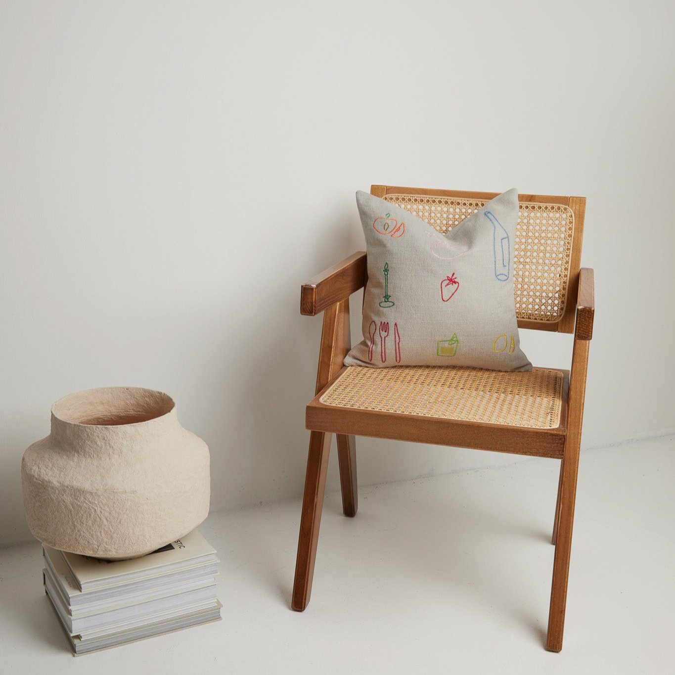 Neutral linen cushion with colourful embroidered icons placed on a cane wooden chair, styled with a paper clay vase on stacked books