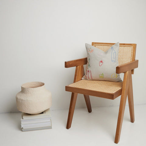 Minimalist styling of embroidered icons cushion on cane wooden chair beside a textured vase.