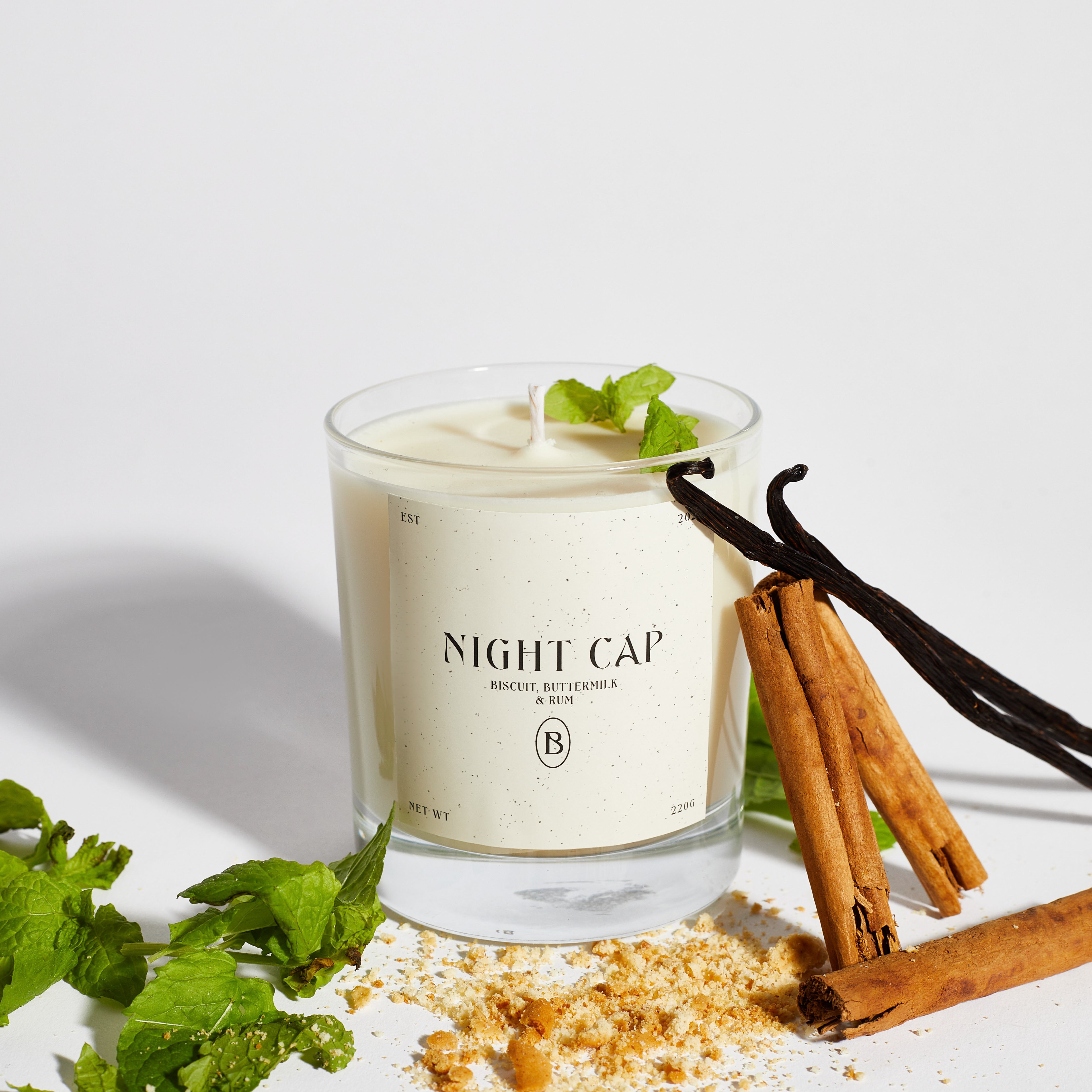 Boujie Night Cap jar candle in a clear glass vessel, surrounded by fresh mint leaves, cinnamon sticks, vanilla pods, and biscuit crumbs, capturing the indulgent scent of biscuit, buttermilk, and rum.