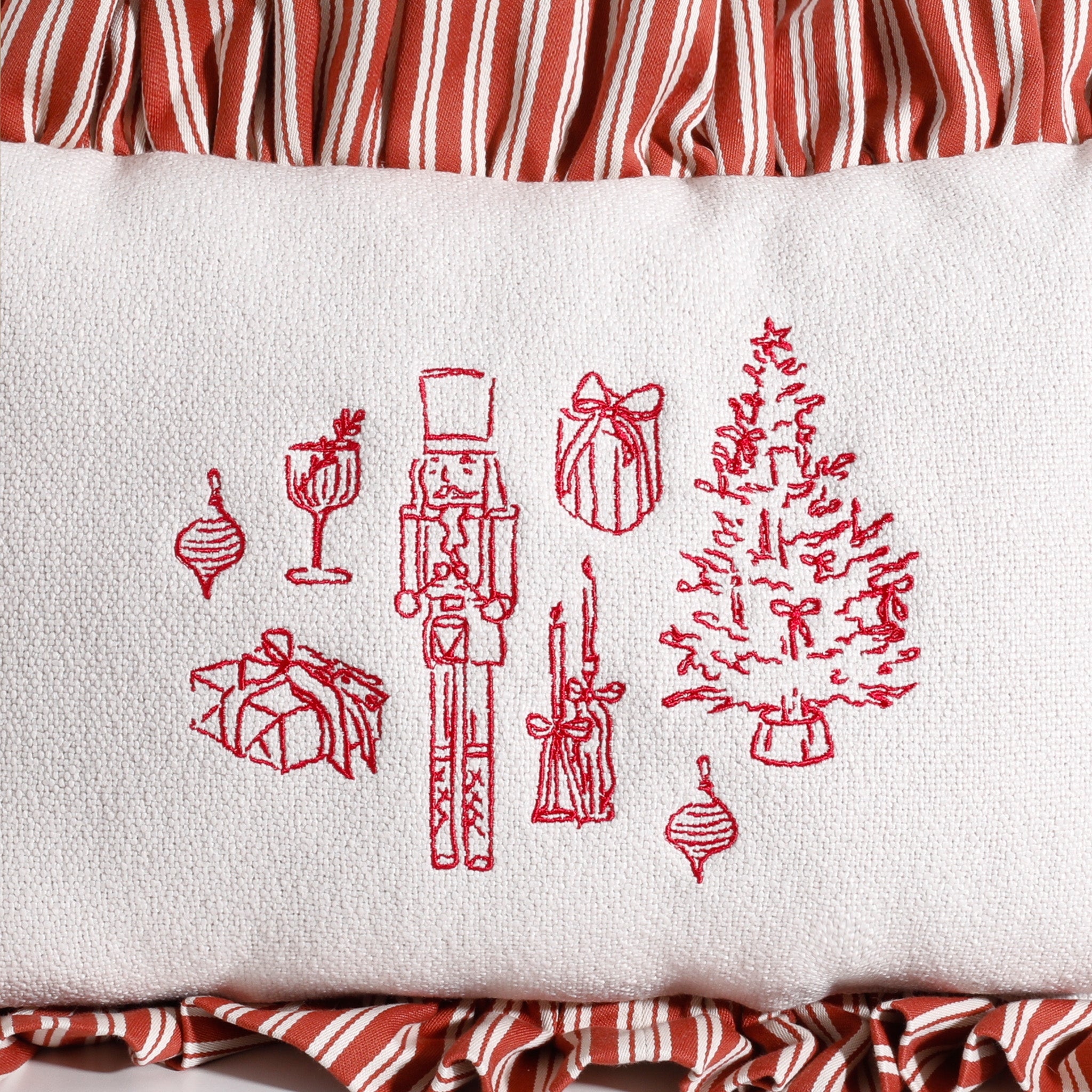 Close-up of red holiday embroidery on a white decorative pillow featuring a nutcracker, Christmas tree, wrapped gifts, ornaments, and candles with a red and white striped ruffle border.
