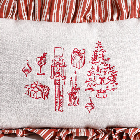 Close-up of red holiday embroidery on a white decorative pillow featuring a nutcracker, Christmas tree, wrapped gifts, ornaments, and candles with a red and white striped ruffle border.