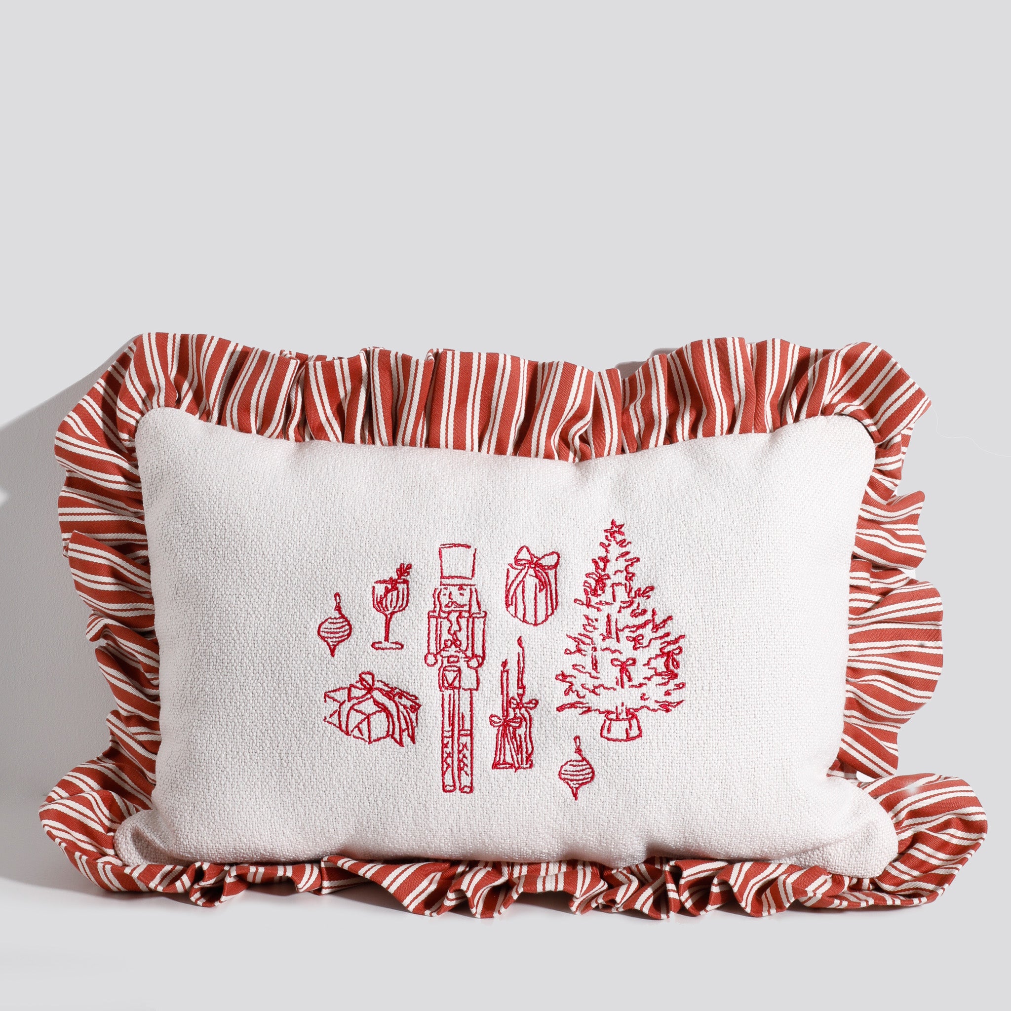 A rectangular white Christmas pillow with red embroidered holiday icons, including a nutcracker, Christmas tree, ornaments, gifts, and candles, surrounded by a red and white striped ruffled edge.