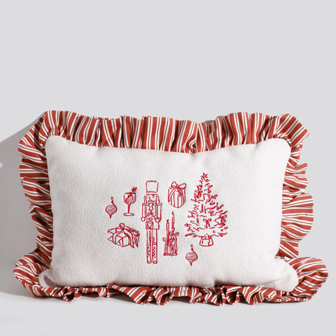A rectangular white Christmas pillow with red embroidered holiday icons, including a nutcracker, Christmas tree, ornaments, gifts, and candles, surrounded by a red and white striped ruffled edge.