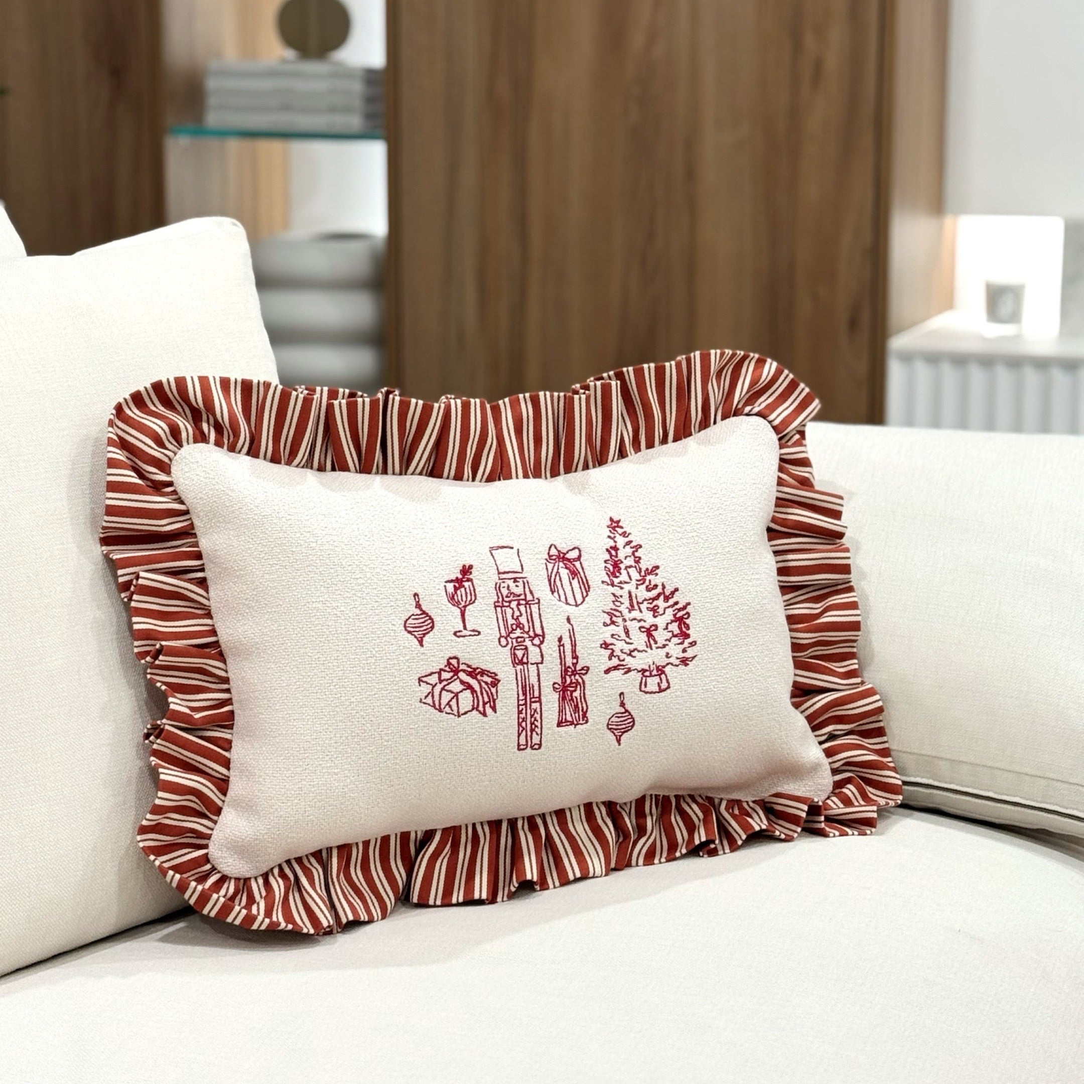 Embrodiered festive line-art burgundy detail on a cream cushion with striped ruffle edging. 