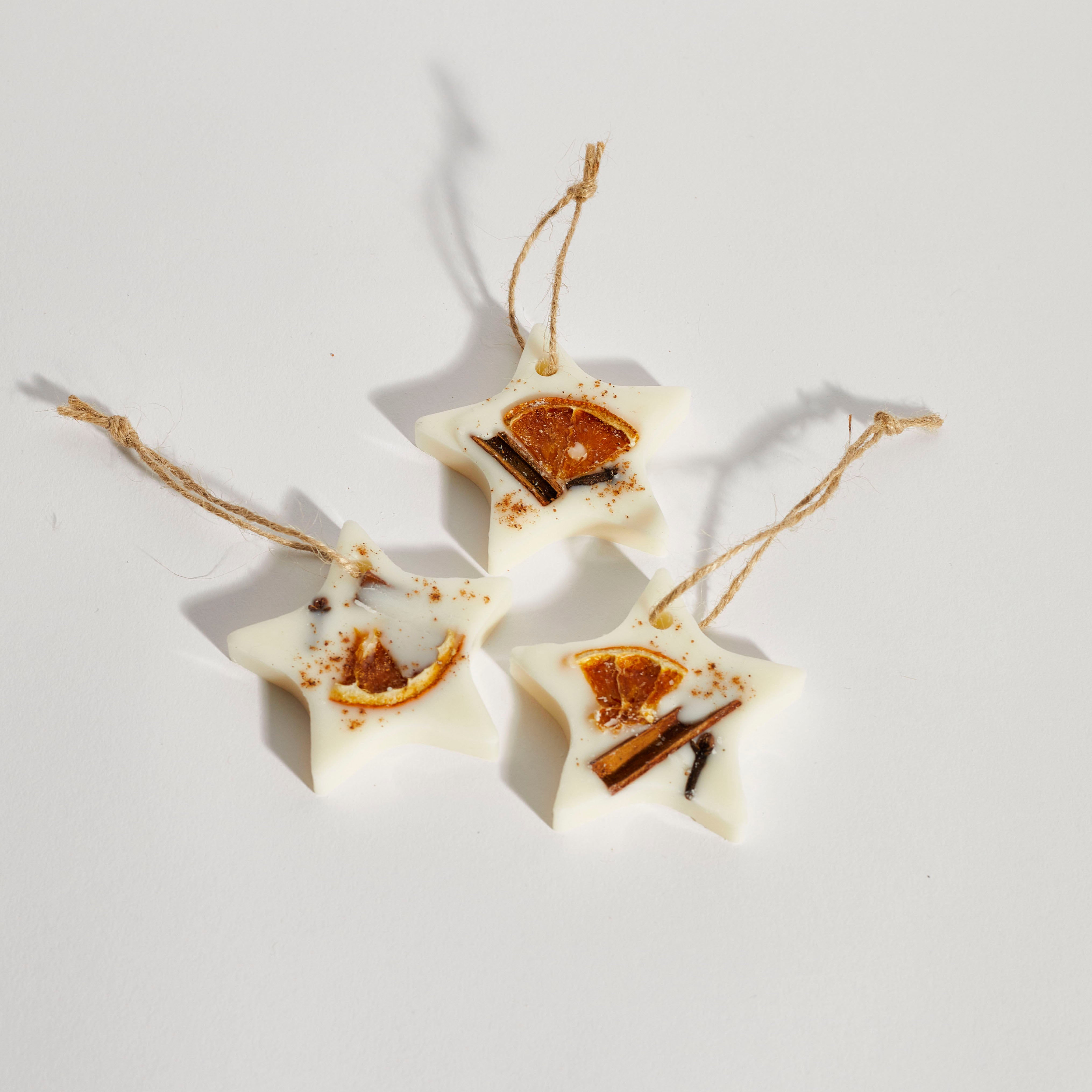 Three white wax tarts with star-shaped patterns, adorned with dried botanicals, displayed against a white background with twine hanging from each.